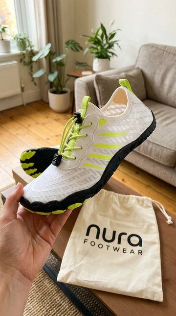 White sneaker with green accents held in a hand, next to a 'nura footwear' drawstring bag in a living room.