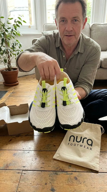 Man holding a pair of white sneakers with neon green laces on a wooden table, with a 'nura footwear' bag in the foreground.