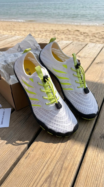 White sneakers with black soles and neon green laces on a wooden surface with a beach background.