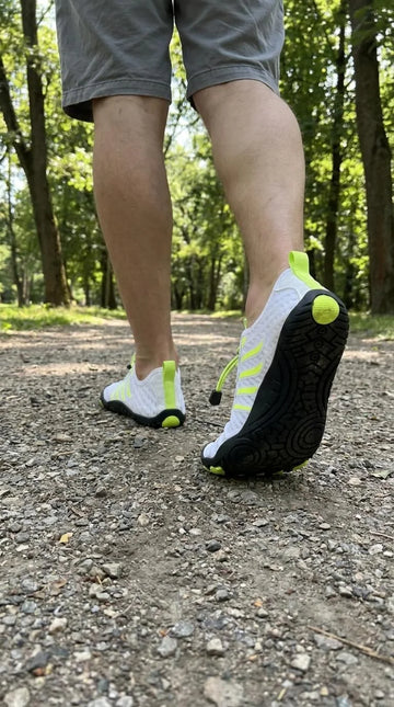 Person wearing white running shoes with black soles on a gravel path in a forest.