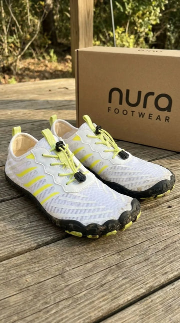 White sneakers with yellow accents and black soles on a wooden surface next to a Nura Footwear box.